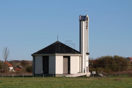 Newly built modern white and grey catholic church tall bell tower with iron cross on top and large ringing bell mechanism in middle surrounded with wire fence and uncut grass with dense trees and urban family houses in background