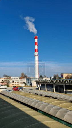 Photo for Renovated newly painted high industrial red and white chimney with multiple cell phone antennas and metal stairs overlooking small power plant building next to parking lot and old marketplace on clear blue sky background - Royalty Free Image
