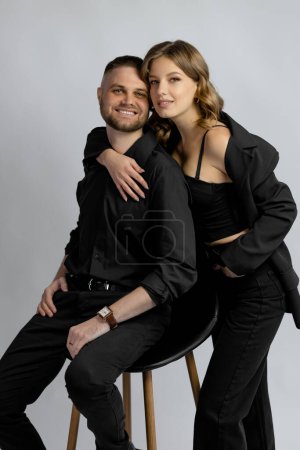 Téléchargez les photos : Une jeune femme et un jeune homme en vêtements noirs se serrent dans leurs bras et sourient assis sur une chaise sur un fond gris. Photo de haute qualité - en image libre de droit