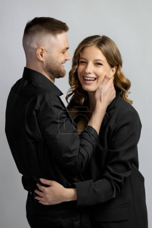 Téléchargez les photos : Gros plan de jeune femme et jeune homme en vêtements noirs debout étreignant et souriant sur fond gris. Photo de haute qualité - en image libre de droit