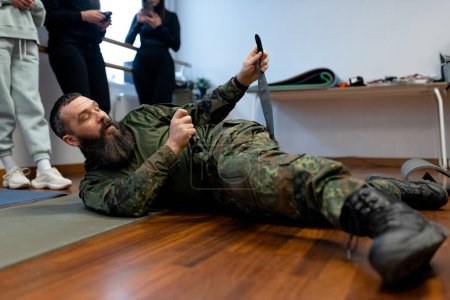 A bearded man in his 40s, a military medic lies on the floor and demonstrates to civilians in the room how to apply a combat medical tourniquet to stop bleeding on a leg. High quality photo