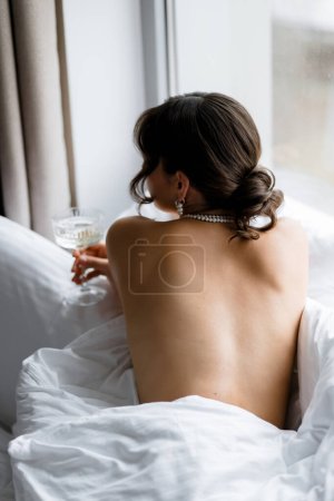 Téléchargez les photos : Une belle mariée brune se couche sans sous-vêtements le matin dans la chambre sur le lit de la chambre avec une coupe de champagne. Photo de derrière. Photo de haute qualité - en image libre de droit