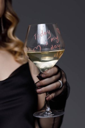 Eine Person im schicken schwarzen Outfit hält ein Glas Weißwein in der Hand. Das Glas trägt eine handschriftliche Botschaft Heute ist mein Tag, symbolisiert einen Moment der persönlichen Feier