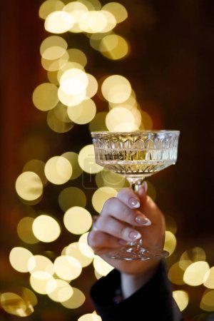 Eine Nahaufnahme einer Hand, die anmutig ein Kristallglas hält, aufgenommen vor dem Hintergrund warmer, goldener Bokeh-Lichter, die eine Atmosphäre der Feier und des Luxus vermitteln