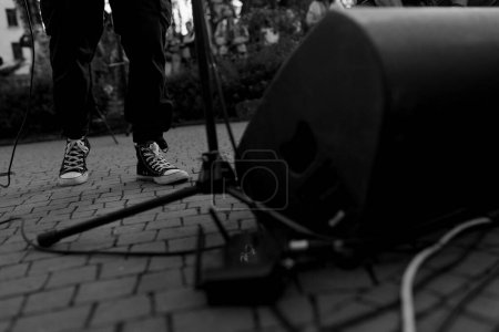 Una imagen en blanco y negro que captura la vista del suelo de unas zapatillas de deporte de músico, un soporte de micrófono y un altavoz sobre una superficie pavimentada, evocando la energía bruta del rendimiento callejero