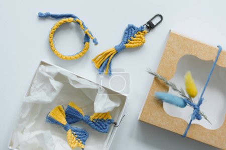 A flat lay of handcrafted macrame accessories in blue and yellow, including a bracelet, keychain, and earrings displayed beside an elegantly wrapped gift box
