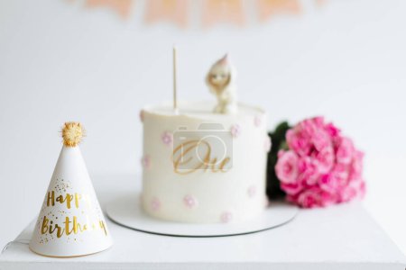 A beautifully decorated first birthday cake adorned with a One inscription and a kitten figurine, complemented by a festive hat and vibrant flowers, capturing the essence of a joyful celebration
