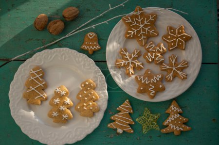 Foto de Pintados tradicionales panes de jengibre de Navidad dispuestos en platos blancos en la antigua mesa pintada vintage a la luz del día, varios árboles de formas de Navidad, estrellas y copos de nieve - Imagen libre de derechos