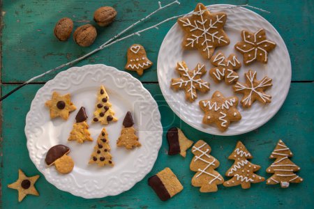 Photo for Painted Christmas gingerbreads and custard cookies arranged on white plates on old vintage painted table in daylight, various xmas shapes trees, stars and snowflakes - Royalty Free Image