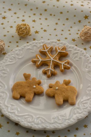 Photo for Painted traditional Christmas gingerbreads arranged on white plate, tablecloth with golden stars, snowflake and reindeer shapes with ball arrangements - Royalty Free Image