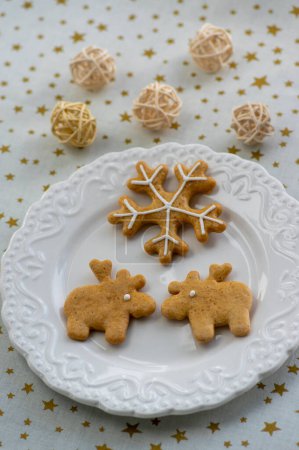 Foto de Panes de jengibre tradicionales de Navidad pintados dispuestos en plato blanco, mantel con estrellas doradas, copos de nieve y formas de renos con arreglos de bolas - Imagen libre de derechos