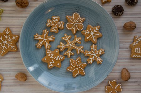 Foto de Pintados tradicionales panes de jengibre de Navidad dispuestos en plato azul sobre mesa de madera blanca a la luz del día, varios árboles de formas de Navidad, estrellas y copos de nieve y botones de chocolate - Imagen libre de derechos