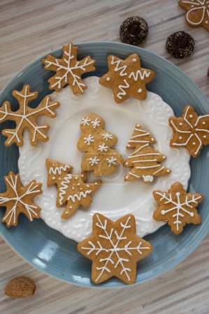 Foto de Pintados tradicionales panes de jengibre de Navidad dispuestos en plato azul sobre mesa de madera blanca a la luz del día, varios árboles de formas de Navidad, estrellas y copos de nieve y botones de chocolate - Imagen libre de derechos