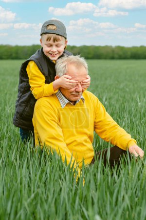 Foto de Family activity outdoors game and playtime positive. The boy closes his eyes to the grandfather sitting on the green grass. High quality photo - Imagen libre de derechos
