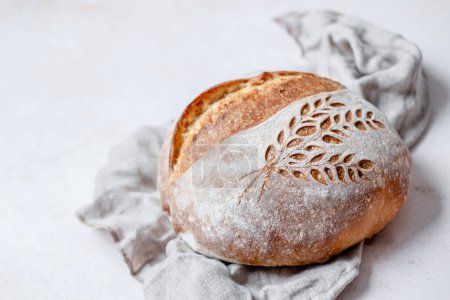 Foto de Rustic Italian Bread with Lievito Madre on a napkin on a white table. Scoring an ears and wheat pattern. Cooking of healthy bread from alternative flours. Fermented food. High quality photo - Imagen libre de derechos