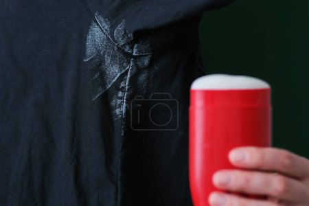 Téléchargez les photos : Teinture désodorisante sur chemise noire. sur le fond une main tenant un déodorant rouge. bâton vie quotidienne tache concept. Photo de haute qualité - en image libre de droit