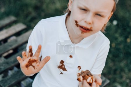 Téléchargez les photos : Un garçon avec les mains et le visage en chocolat. Brun taches de nourriture sur les vêtements blancs. à l'extérieur. Photo de haute qualité - en image libre de droit