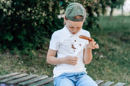 Foto de Un niño mostrando manchas de chocolate en su ropa sentado en un banco de madera en el parque. al aire libre. El concepto de limpieza de manchas en la ropa. Foto de alta calidad - Imagen libre de derechos