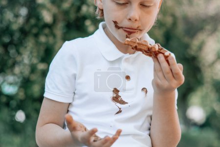 Téléchargez les photos : Un enfant mange un gâteau au chocolat assis sur un banc en bois dans le parc, le visage et la main dans du chocolat fondu. Des taches de chocolat sale sur des vêtements blancs. à l'extérieur. Photo de haute qualité - en image libre de droit