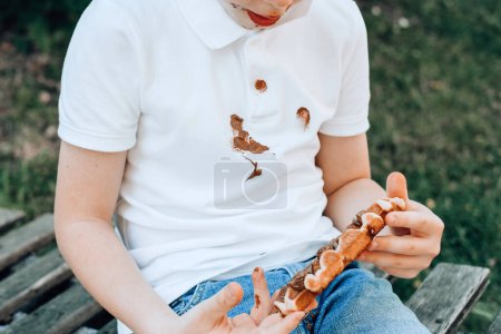 Téléchargez les photos : Taches de chocolat sur les vêtements blancs. Un garçon qui mange des gaufres belges avec des mains collantes en chocolat. Des vêtements abîmés. à l'extérieur. Photo de haute qualité - en image libre de droit