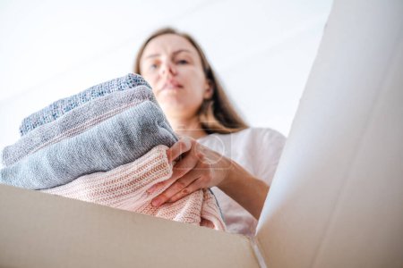Photo for Low angle view of a woman sorting clothes and putting stack of knitted sweaters in a box for selling, donation or recycling. Conscious and environmentally consumption. High quality photo - Royalty Free Image