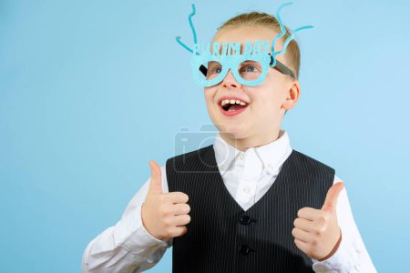 Foto de Divertido niño en un traje elegante con gafas de día Purim, mostrando su mano super, sonríe el fondo azul. Concepto de celebración Purim. Fiesta de carnaval judío. Foto de alta calidad - Imagen libre de derechos