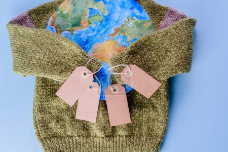 Kreative Konzepte kaufen weniger und übermäßigen Konsum. Die Pullover-Ärmel halten die Tags in der Umarmung des Planeten auf blauem Hintergrund. Verantwortungsvoller Konsum. Ansicht von oben. Hochwertiges Foto
