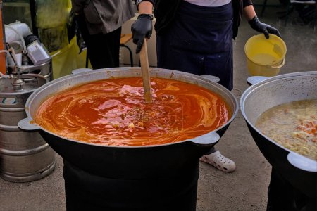 Foto de Comida sana callejera para llevar - borscht rojo con pimientos en un caldero grande. El cocinero pone una porción de borscht en el recipiente. Deliciosa comida en oferta en la feria de comida callejera, evento, festival. Ucrania. - Imagen libre de derechos