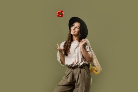 Photo for Vegetarian woman wear casual clothes and black hat hold net eco bag with vegetables and throw up a red pepper in air - Royalty Free Image