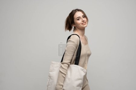 Photo for Brunette cheerful millennial woman holding white eco bag standing over white studio background. Lady holding flax shopper handbag. Fashion and ecology concept - Royalty Free Image