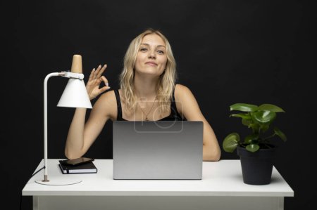 Téléchargez les photos : Femme d'affaires blonde joyeuse en chemise noire montrant le geste OK tout en travaillant sur ordinateur portable sur fond de studio bleu. Femme entrepreneure heureuse faisant signe d'approbation, exprimant la positivité - en image libre de droit