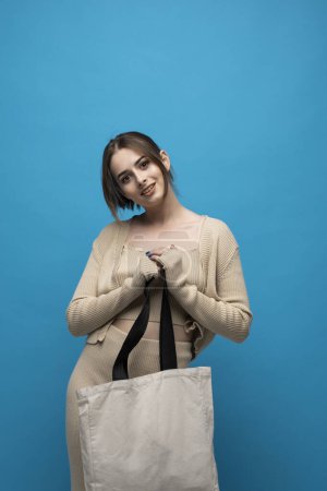 Photo for Brunette cheerful millennial woman holding white eco bag standing over white studio background. Lady holding flax shopper handbag. Fashion and ecology concept - Royalty Free Image