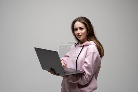 Téléchargez les photos : Portrait de belle jolie fille brune joyeuse dans un sweat à capuche rose tenant dans les mains ordinateur portable créant présentation isolée sur fond gris - en image libre de droit