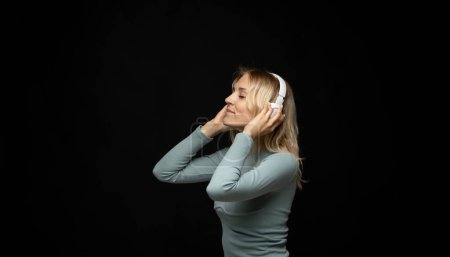 Téléchargez les photos : Joyeux jeune femme blonde fille écouter de la musique dans des écouteurs blancs et se détendre sur fond noir. Charmante fille s'amuser et profiter de la vie. Attrayant femelle profiter de sa chanson préférée - en image libre de droit