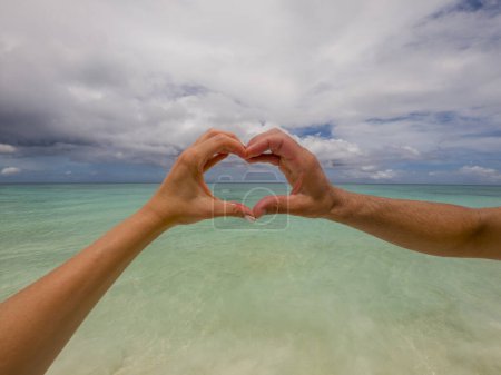 Téléchargez les photos : Couple formant un cœur avec leurs mains devant la mer verte des Caraïbes et le ciel bleu - en image libre de droit
