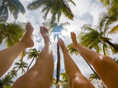 Téléchargez les photos : Jeune couple avec les jambes pointées vers le ciel bleu avec des palmiers et des nuages - en image libre de droit