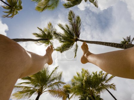 Téléchargez les photos : Jeune homme avec les jambes pointant vers le ciel bleu avec des palmiers et des nuages - en image libre de droit