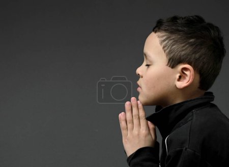 Téléchargez les photos : Petit garçon priant dieu sur fond sombre en studio - en image libre de droit