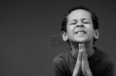 Téléchargez les photos : Portrait de petit garçon priant Dieu sur fond sombre en studio, noir et blanc - en image libre de droit