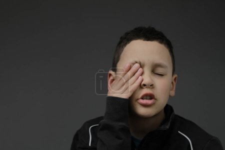 Téléchargez les photos : Petit garçon se sentant somnolent et frottant les yeux avec les mains sur fond sombre - en image libre de droit