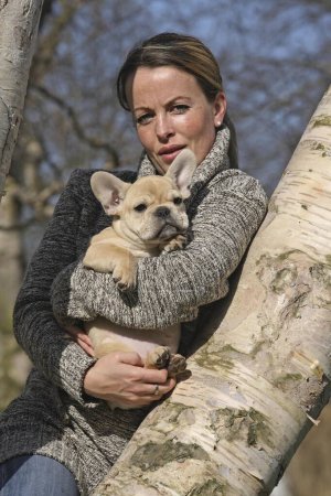 Junge blonde Frau lehnt an einer Birke und hält eine 12 Wochen alte französische Bulldogge im Arm Weitere Motive zu diesem Thema sind auf Anfrage erhältlich