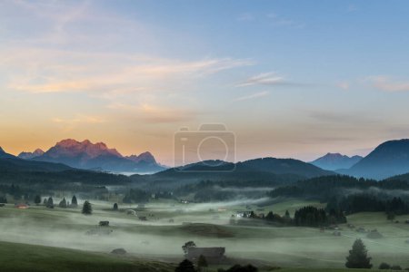 Bergwiesen mit kleinen Hütten im leichten Bodennebel und Zugspitzmassiv im Hintergrund bei Sonnenaufgang, Krn, Bayern, Deutschland, Europa 