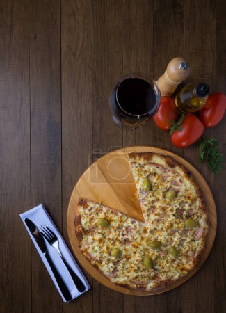 Foto de Gratin Sirloin Pizza servida sobre tabla de madera. Mozzarella, solomillo canadiense, cuajada cremosa y aceitunas verdes. Rebanada servida. Copa de vino. - Imagen libre de derechos