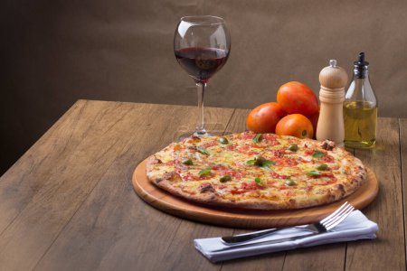 Photo for Pizza Marguerita made with tasty pizza dough, Mozzarella, tomatoes, marjoram and green olives. On the wooden board. Napolitan Pizza. Glass of Red Wine, olive oil, pepper grinder, fork and knife. Gourmet gastronomy. - Royalty Free Image