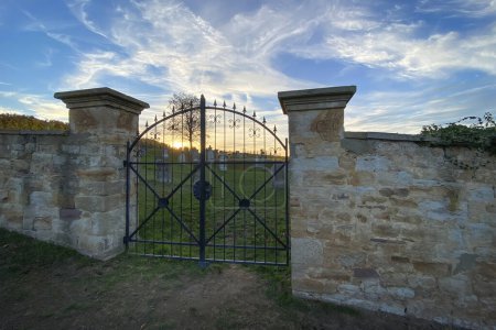 Téléchargez les photos : Porte de fer à l'entrée du cimetière avec de vieilles pierres tombales près de Flonheim, Rhin Hesse, Allemagne ciel bleu avec nuages au coucher du soleil - en image libre de droit