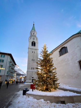 Photo for Cortina d'Ampezzo, Italy - January 26, 2023: Illuminated Christmas tree with Catholic Church Santi Filippo e Giacomo Apostoli in winter against blue sky - Royalty Free Image