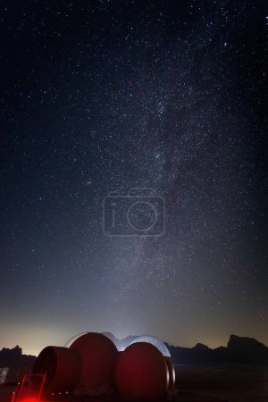 Ciel nocturne plein d'étoiles et de la voie lactée au-dessus du désert de Wadi Rum avec vue partielle sur une tente à bulles et un paysage rocheux