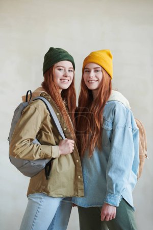 Vertikales mittellanges Studioporträt von zwei rothaarigen jungen kaukasischen Zwillingen, die stilvolle lässige Outfits mit Rucksäcken tragen und in die Kamera lächeln