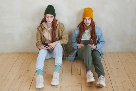 Photo for High angle view studio portrait of young twin sisters wearing fashionable casual clothes sitting on wooden floor using gadgets - Royalty Free Image