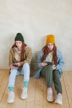 Photo for High angle view of stylish twin sisters wearing casual clothes sitting on floor surfing Internet on their gadgets - Royalty Free Image